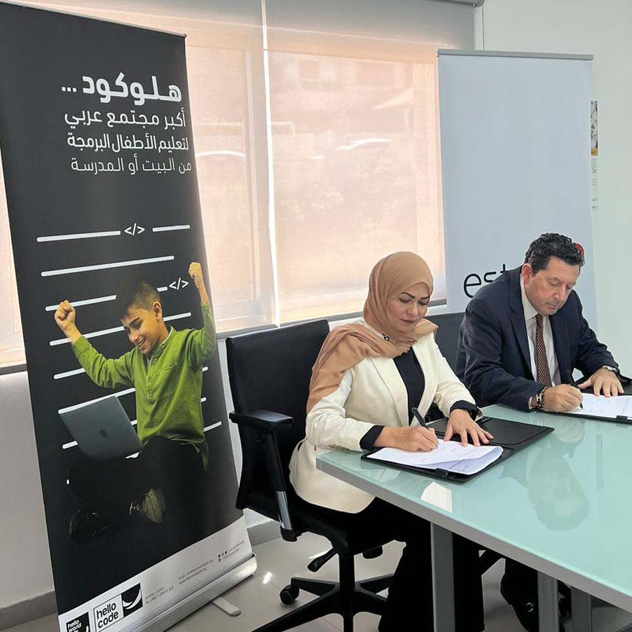 A woman and a man are seated at a table, signing documents. Behind them is a banner with Arabic text and an image of a boy celebrating with raised fists. The woman is wearing a headscarf, and the man is in a suit. They are in an office setting with large windows.