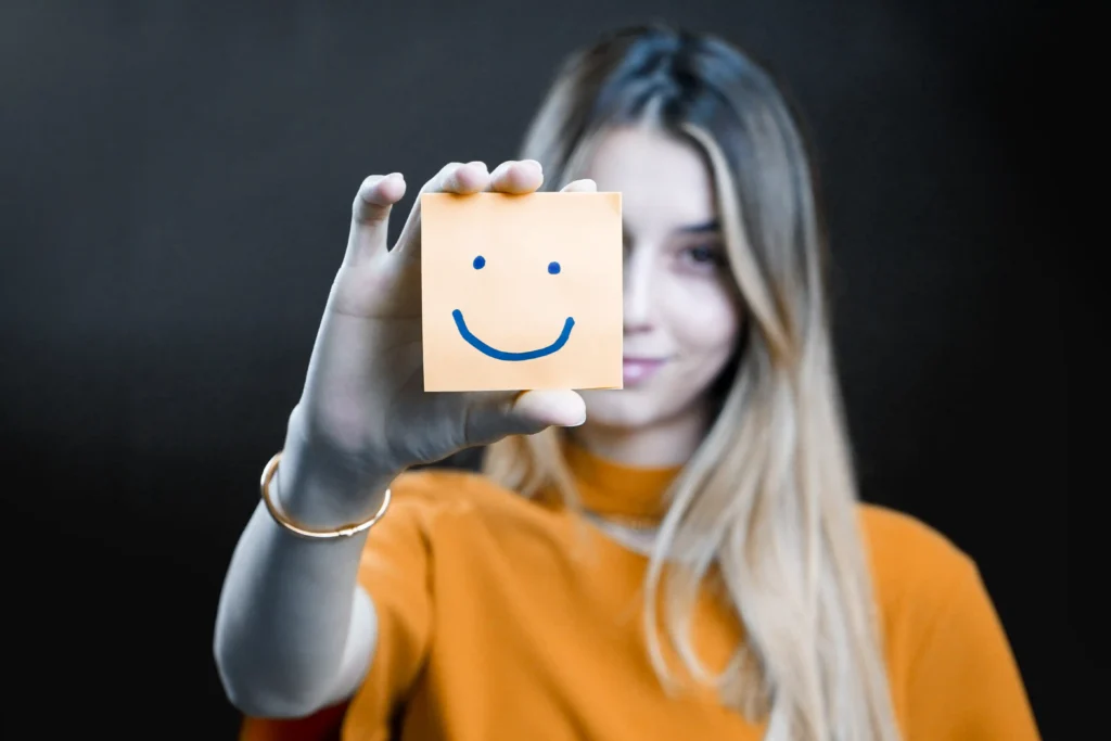 A person with long hair holds a sticky note in front of their face, featuring a simple smiley face. Wearing a yellowish-orange shirt, they stand against a dark background. Only their eyes and part of the face are visible, giving the impression they're offering an unexpected moment of joy amidst everyday services.
