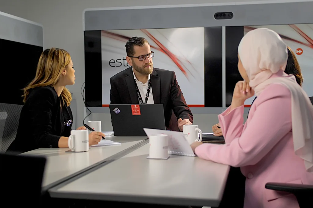 A group of people sit around a conference table in a modern office. A man with glasses and a beard is discussing various services, while two women listen attentively. Laptops, papers, and coffee mugs are on the table. A large screen displaying a presentation is in the background.