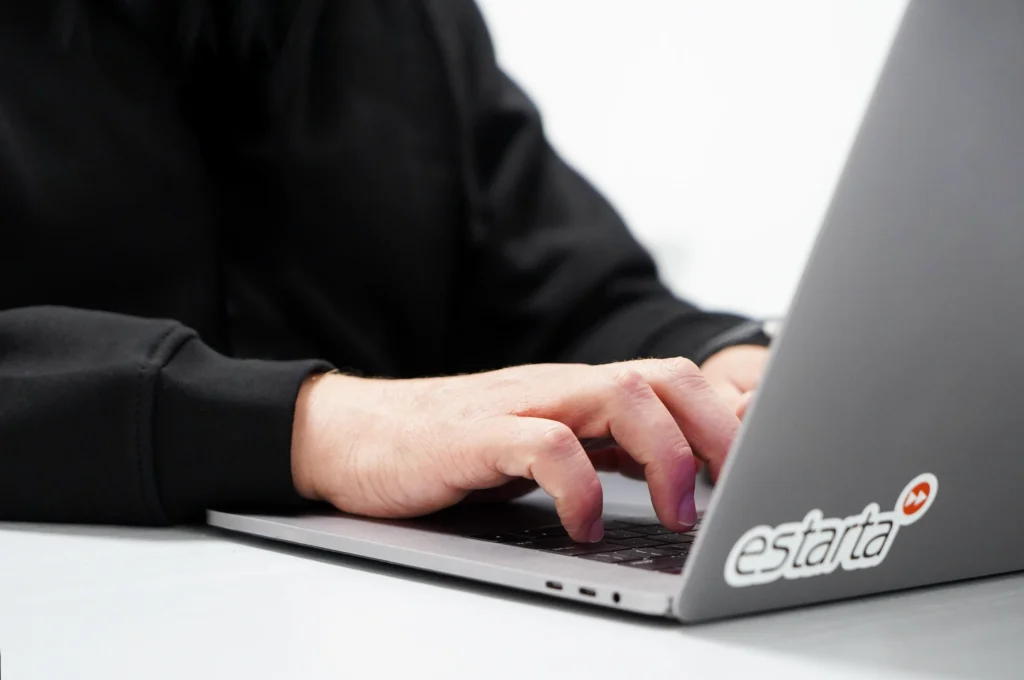 Close-up of a person typing on a laptop with an "estarta" sticker. The person, wearing a long-sleeve black shirt, has their hands and part of their arms visible. The plain white background focuses attention on the scene, hinting at professional services.