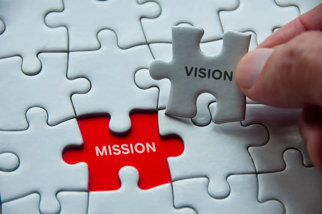 A hand places a puzzle piece labeled "VISION" into an almost complete puzzle, leaving one red piece labeled "MISSION" exposed. The white puzzle pieces symbolize the Vision and Mission of Estarta in achieving a goal.