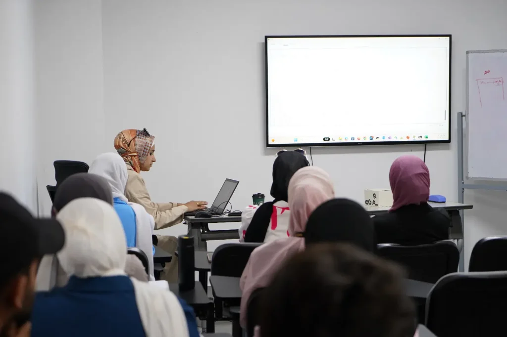 A group of people seated in a classroom are facing a presentation screen. A woman at the front, wearing a hijab, is working on a laptop connected to the screen. The audience includes individuals with hijabs, watching attentively. The whiteboard on the right lists points from the Networking Course hosted by Estarta.