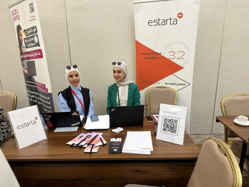 Two women wearing hijabs sit behind a table with laptops and promotional materials at a job fair. A banner behind them reads "estarta" and mentions "32 Years in the Industry" and "1500+ Employees." Chairs surround the table, and an EFE QR code is visible on a stand.