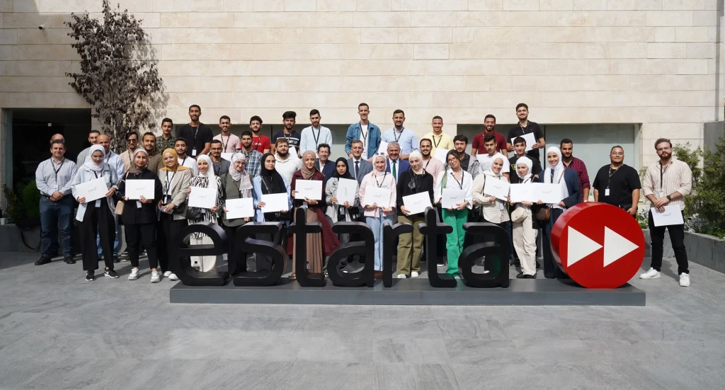 A large group of people standing and posing for a photo in front of a large sign that reads "eSTARTA". They are holding certificates and wearing badges, indicating a formal graduation ceremony, possibly linked to UJ Training. The backdrop is a beige stone wall.